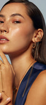 Woman wearing gold jewelry including earrings, necklace, and ring against a neutral background